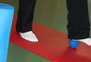 Eine Person steht auf einer Sportmatte und bewegt mit dem Fuß einen kleinen Ball. Daneben steht eine Faszienrolle.