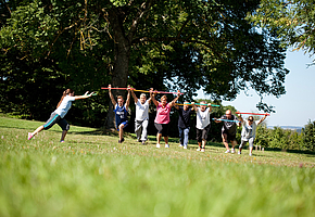Eine Gruppe von Patientinnen und Patienten machen im Park Gymnastikübungen unter der Anleitung einer Physiotherapeutin.