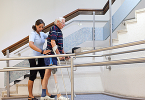 Therapeutin und Patient mit Beinprothese und Krücken gehen gemeinsam im Therapiebereich.